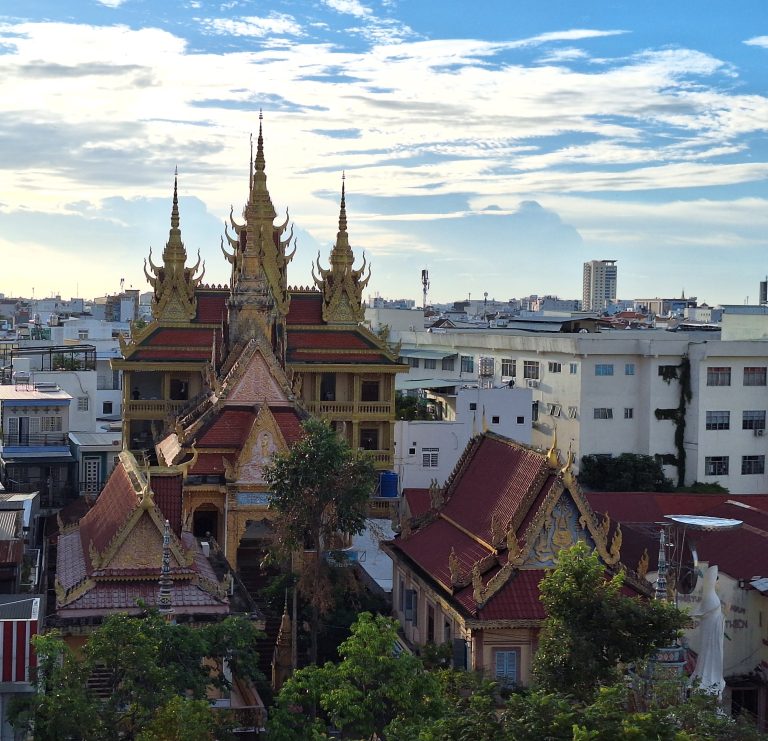 Góc chụp từ trên cao xuống khuôn viên ngôi chùa Khmer Munir Ansay độc đáo tại Cần Thơ, làm nổi bật những mái tháp nhọn màu vàng rực rỡ mang đậm nét kiến trúc Angkor truyền thống, bao quanh là rặng cây xanh và các tòa nhà đô thị.
