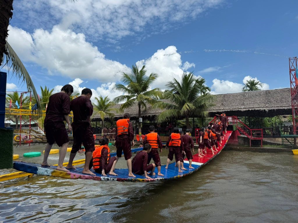 Nhóm du khách mặc áo bà ba và áo phao đang tham gia trò chơi tập thể teambuilding trên cầu phao lắc lư giữa hồ nước tại Làng du lịch sinh thái Ông Đề Cần Thơ.