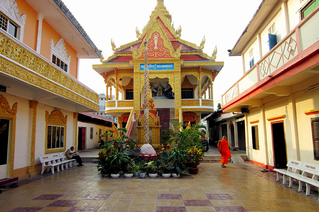Góc nhìn từ sân trong hướng về tòa chính điện hai tầng màu vàng rực rỡ của chùa Munir Ansay Cần Thơ, có cột cờ hình lá sen ở giữa sân và bóng dáng một vị sư mặc áo cà sa đang đi lại.