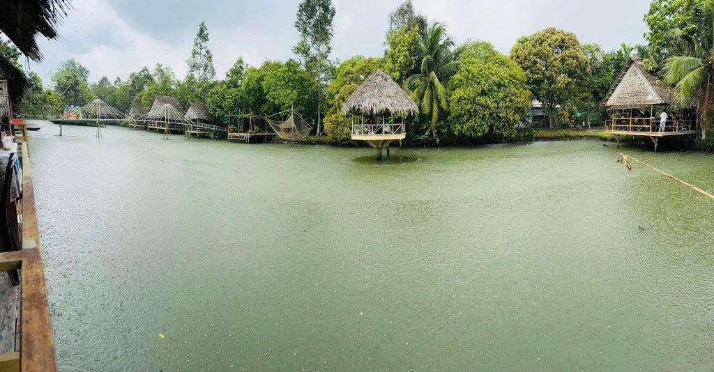 Toàn cảnh hồ nước xanh mát tại khu du lịch sinh thái Lung Cột Cầu Cần Thơ với các chòi lá, cầu khỉ và vó đánh cá đặc trưng miền Tây dưới làn mưa nhẹ.