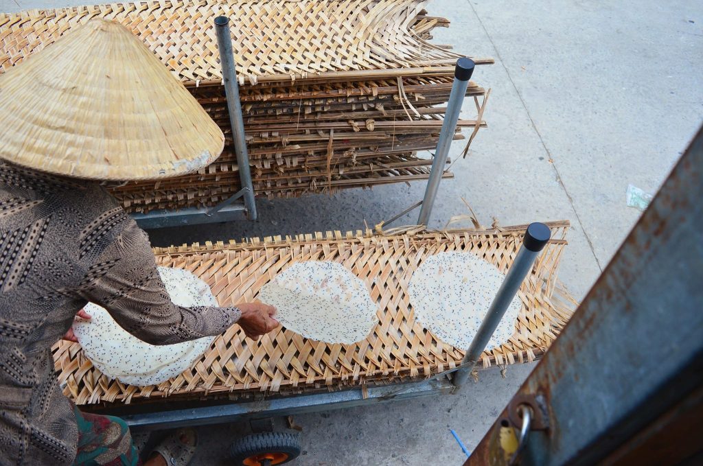 Cận cảnh một người phụ nữ đội nón lá đang xếp bánh tráng mè đen chưa phơi lên vỉ tre đan tại làng nghề bánh tráng Thuận Hưng, Thốt Nốt, Cần Thơ.