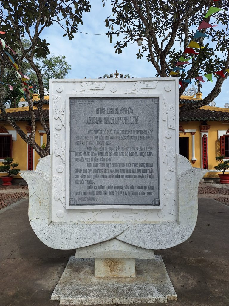 Tấm bia đá xám khắc thông tin lịch sử của Đình Bình Thủy, đặt trên bệ đỡ màu trắng hình hoa sen cách điệu giữa sân đình.