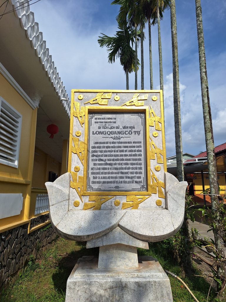 Tấm bia đá khắc thông tin lịch sử của chùa Long Quang Cổ Tự, đặt trên bệ đỡ hình hoa sen màu trắng viền vàng bên cạnh hàng cau cao vút.