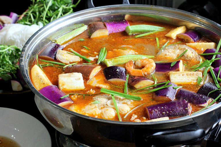 Cận cảnh nồi lẩu mắm Cần Thơ đang sôi trên bếp với đầy đủ nguyên liệu như cà tím, đậu bắp, tôm tươi, cá, thịt và rau nhúng ăn kèm bên cạnh.