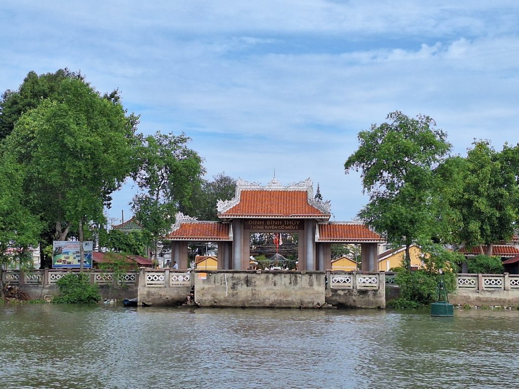 Mặt sau của kiến trúc cổng Đình Bình Thủy với nền móng kiên cố và hàng rào bê tông nhìn từ góc chụp lênh đênh trên mặt nước sông.