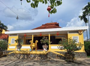 Tòa chính điện chùa Long Quang Cần Thơ với tường sơn màu vàng nổi bật, mái ngói đỏ và hàng hiên viền trắng răng cưa, phía trước bày trí lư hương đồng cùng các chậu cây kiểng bonsai.