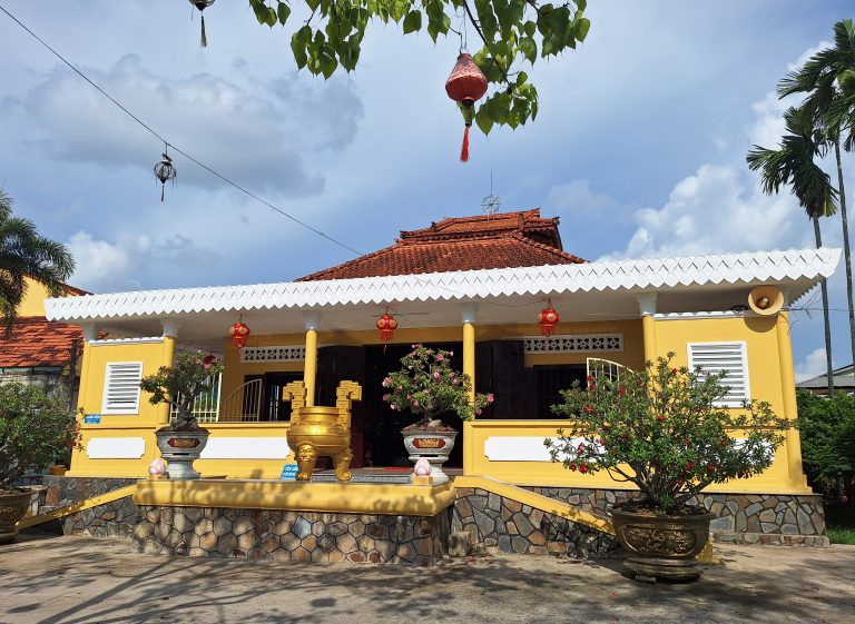 Tòa chính điện chùa Long Quang Cần Thơ với tường sơn màu vàng nổi bật, mái ngói đỏ và hàng hiên viền trắng răng cưa, phía trước bày trí lư hương đồng cùng các chậu cây kiểng bonsai.