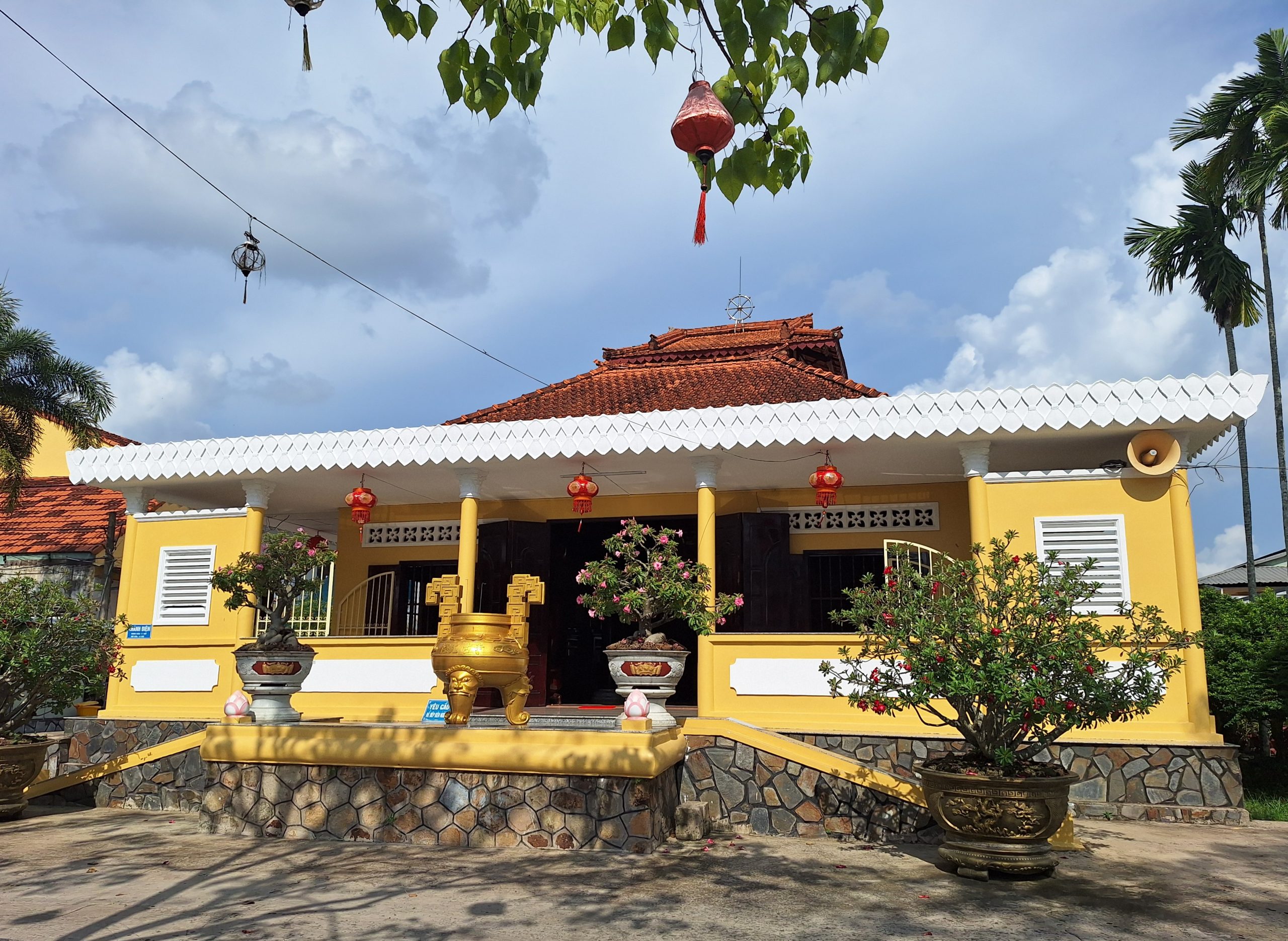 Tòa chính điện chùa Long Quang Cần Thơ với tường sơn màu vàng nổi bật, mái ngói đỏ và hàng hiên viền trắng răng cưa, phía trước bày trí lư hương đồng cùng các chậu cây kiểng bonsai.